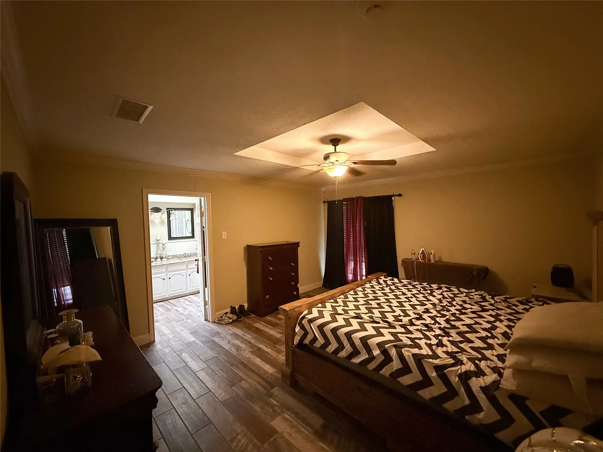Tray ceiling with a ceiling fan, crown molding, wood-finish flooring, and an attached bathroom featuring a dual vanity with a stone countertop