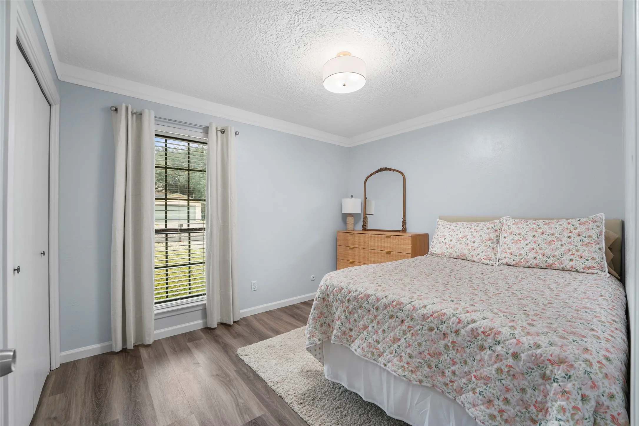 Light-filled room featuring wood-finish flooring, a window with blinds and drapes, crown molding, and a flush-mount ceiling light fixture
