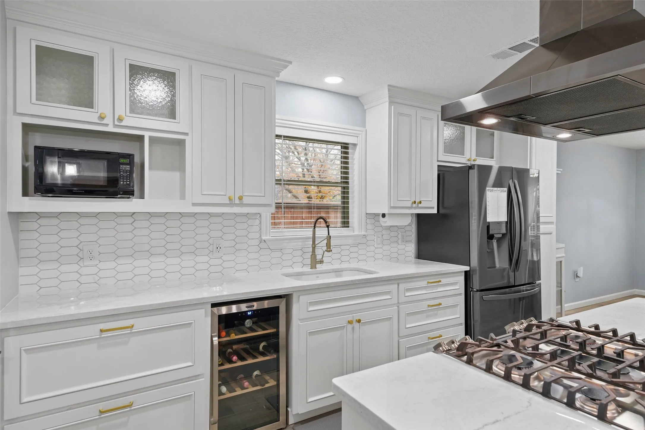 White cabinetry with gold-tone hardware, hexagonal tile backsplash, built-in wine refrigerator, under-mount sink with gooseneck faucet, and countertop gas range