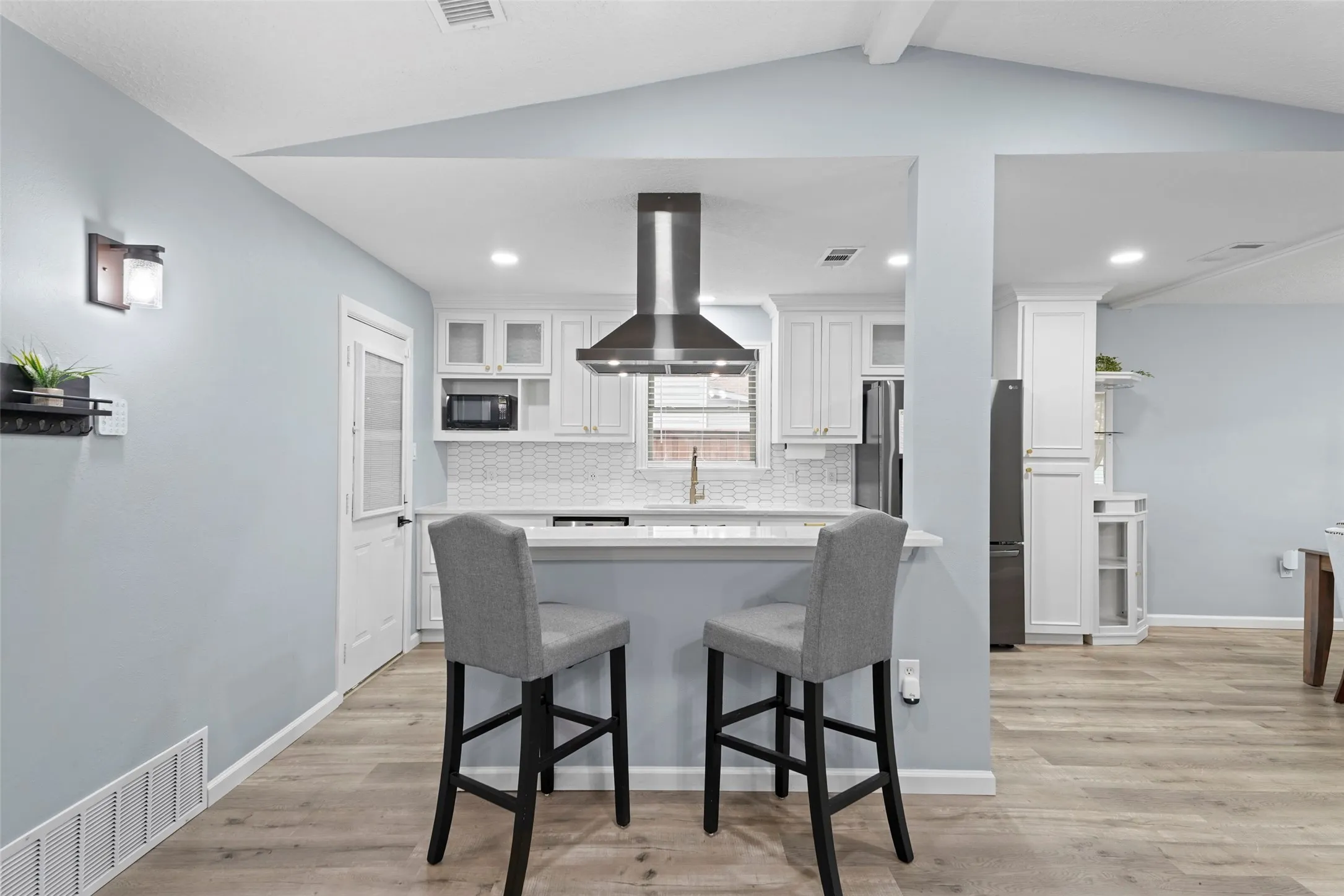 Open-concept kitchen featuring white shaker cabinetry, an island with a breakfast bar, a stainless steel range hood, a white tile backsplash, and wood-finish flooring