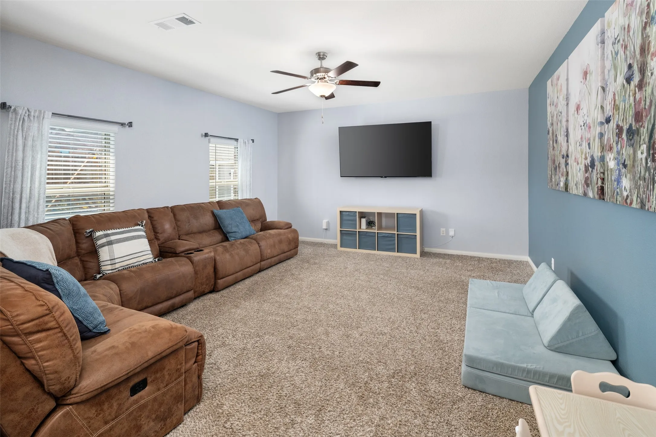 Spacious living area featuring carpet flooring, two windows with blinds, and a ceiling fan with integrated lighting