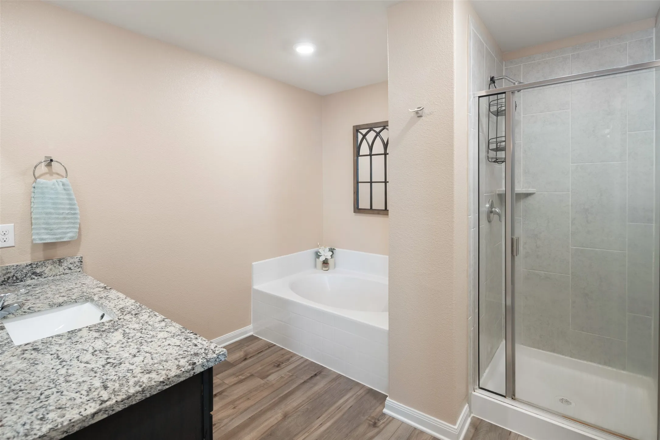 Spa-inspired primary bathroom featuring a soaking tub with white finish, granite speckled countertops, wood-finish flooring, and a frameless glass-enclosed shower with chrome fixtures and tile surround