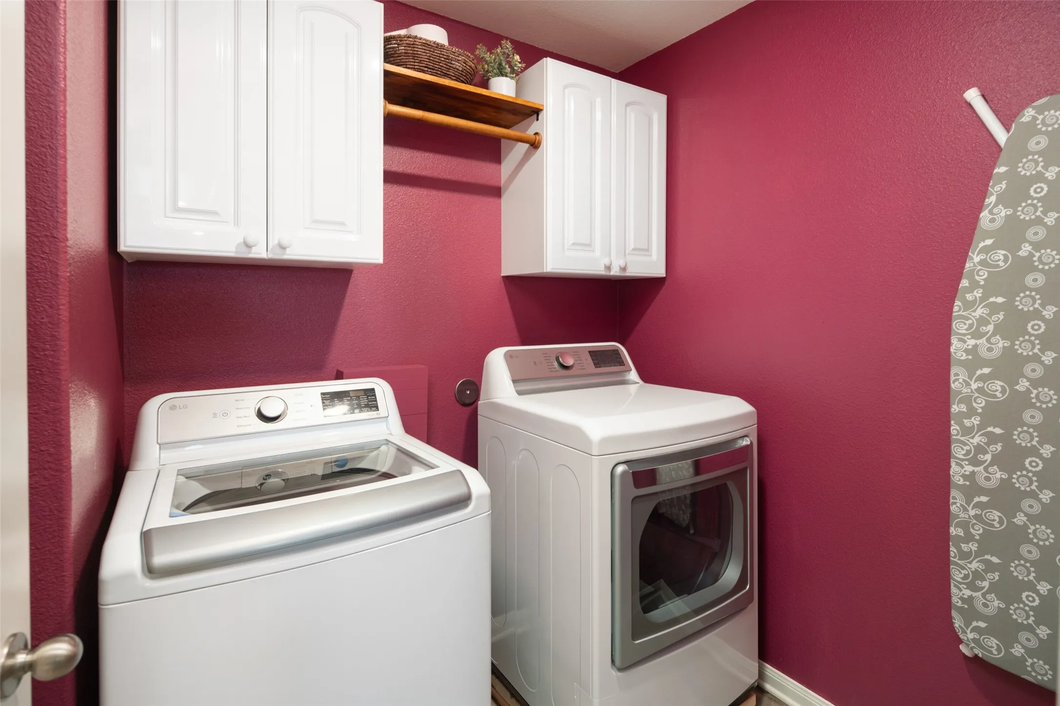 Dedicated laundry room featuring side-by-side white appliances, white cabinetry with upper storage, warm burgundy walls, and wooden shelving for added functionality and organization
