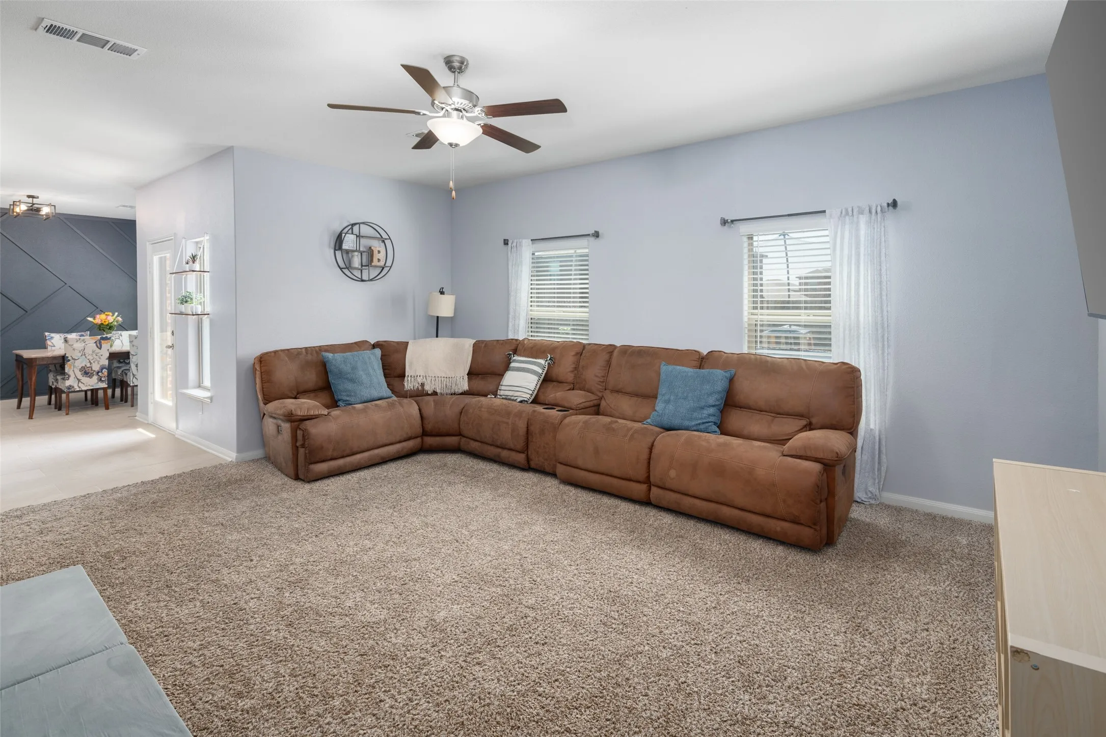 Carpeted living area featuring a ceiling fan with integrated lighting, two windows, and an adjacent dining area with wood-finish flooring and a decorative accent wall