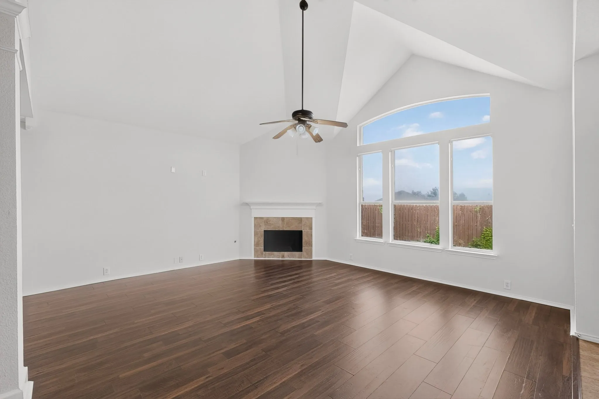 Vaulted ceiling with a ceiling fan, fireplace with a tile surround and mantel, wood-finish flooring, and large windows with an arched transom