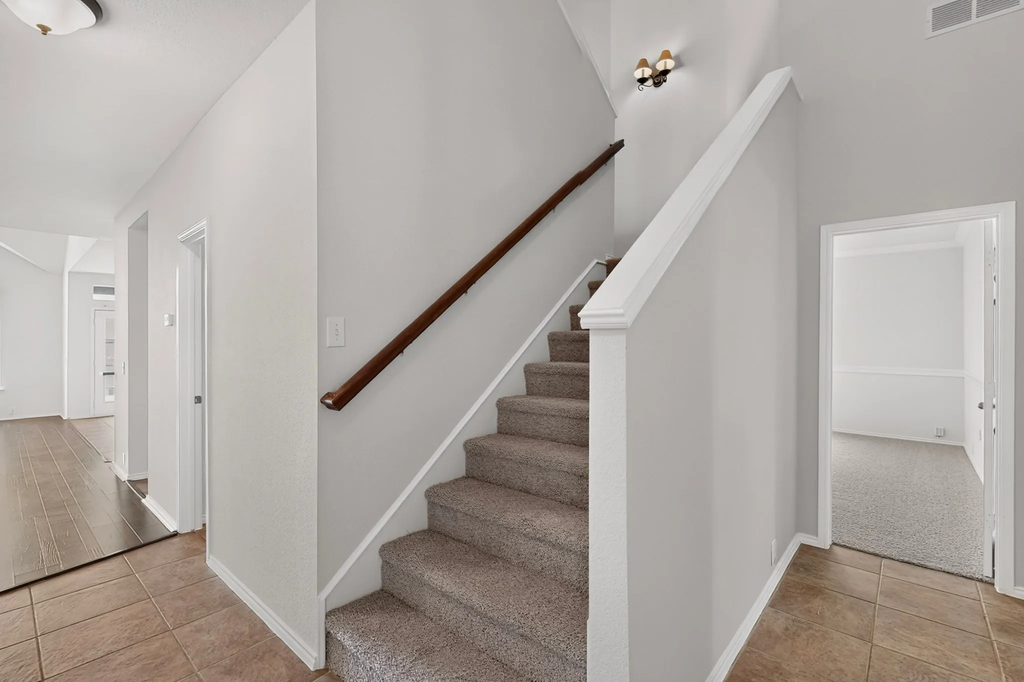 Carpeted staircase with dark wood-finish handrail, wall-mounted light fixture, and tile flooring
