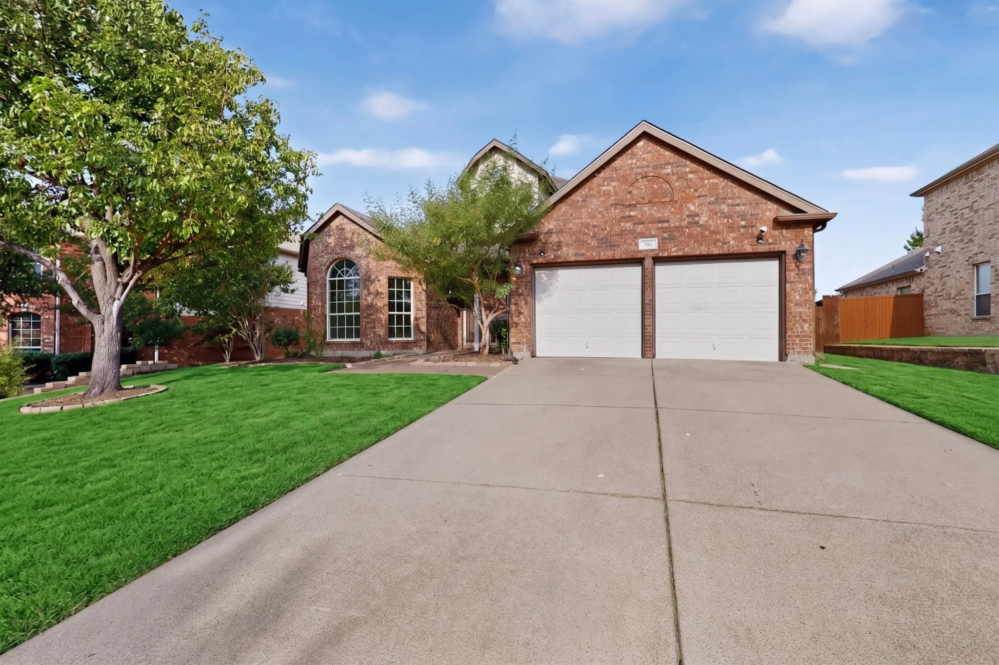 Red brick exterior, arched window, two-car garage, concrete driveway, and a mature tree