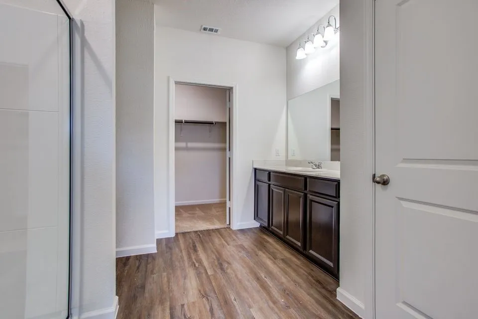Walk-in shower with glass enclosure, dark wood-finish vanity with countertop, wall-mounted mirror, overhead vanity lighting, wood-finish flooring