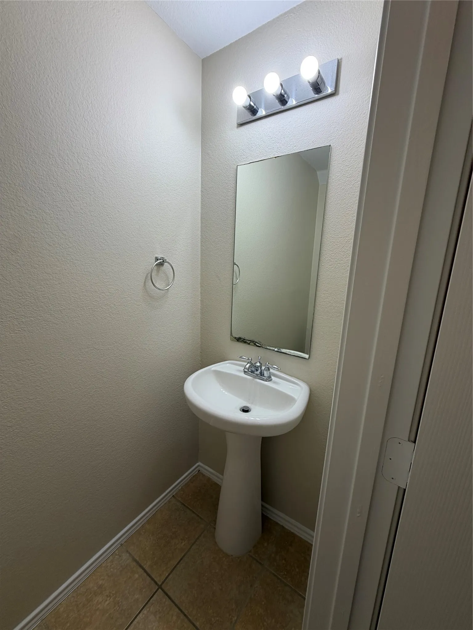 Powder room featuring a pedestal sink with chrome faucet, framed mirror with integrated vanity lighting, tile flooring, and neutral wall finishes