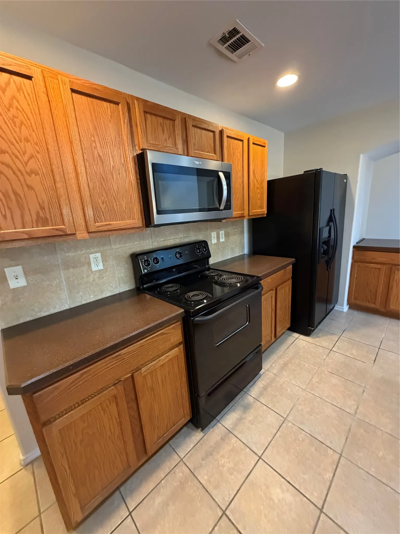 Kitchen featuring wood cabinetry, a stainless steel microwave, black electric range, tile backsplash, and tile flooring
