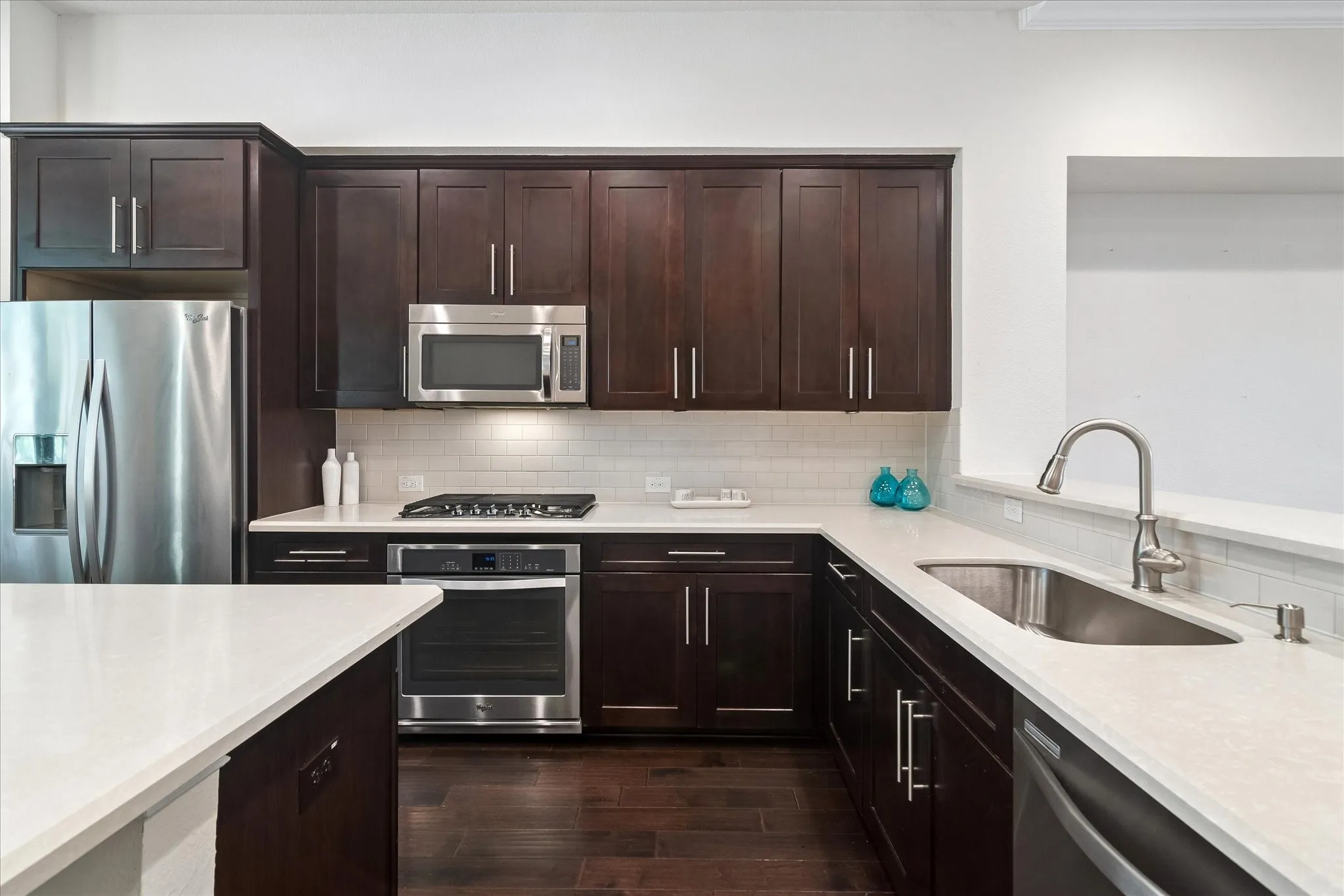 Rich wood-finish cabinetry, stainless steel appliances, white subway tile backsplash, light-toned countertops, and wood-finish flooring