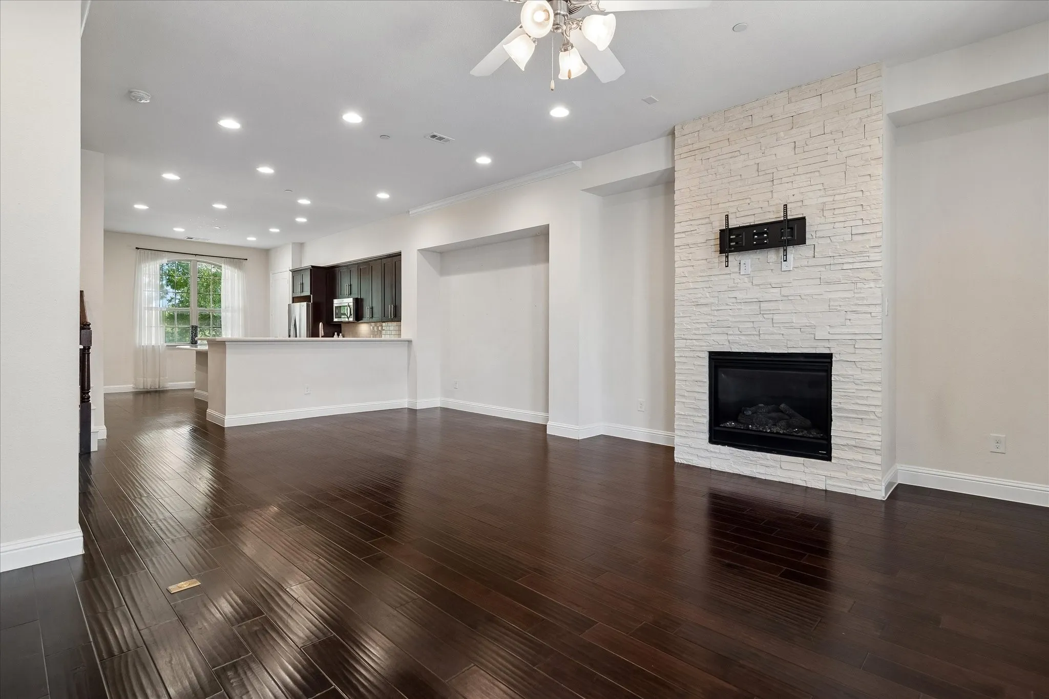 Wood-finish flooring, white stack-stone fireplace, recessed lighting, ceiling fan, and built-in kitchen cabinetry with stainless steel appliances