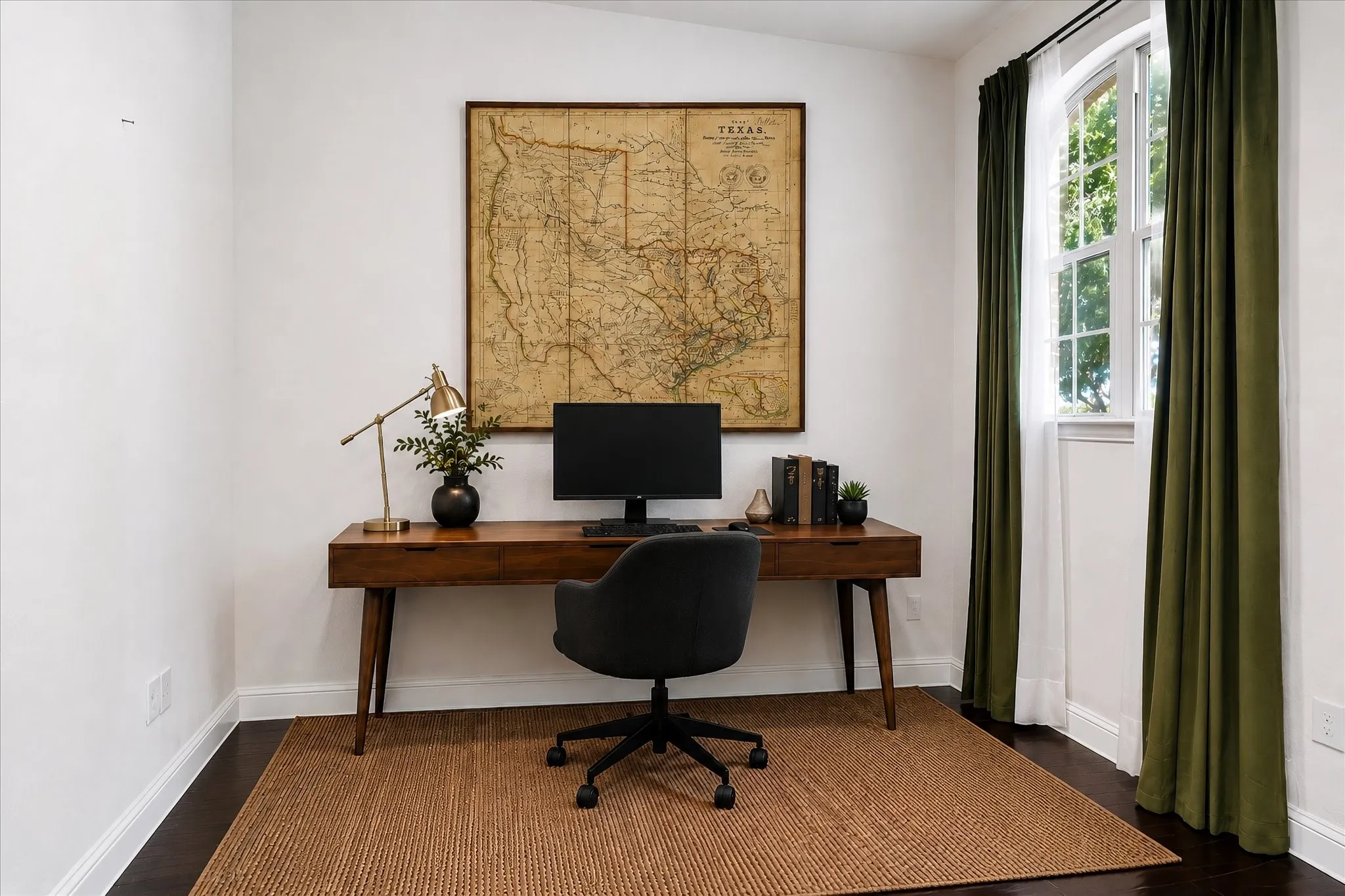 Dedicated home office featuring a mid-century modern wood desk, black task chair, and natural light from a full-height glass door with olive green drapery