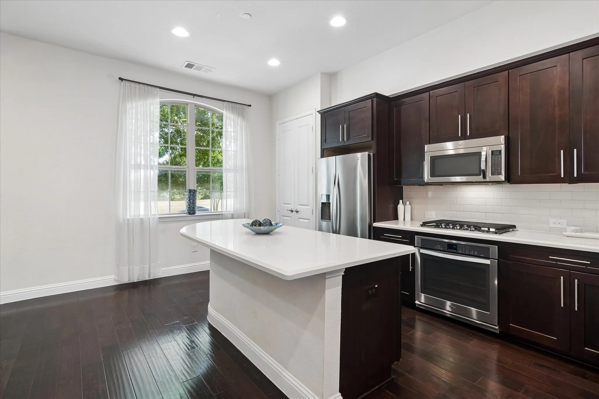 Kitchen featuring dark wood-finish cabinetry, stainless steel appliances, a white subway tile backsplash, and a central island with a white countertop