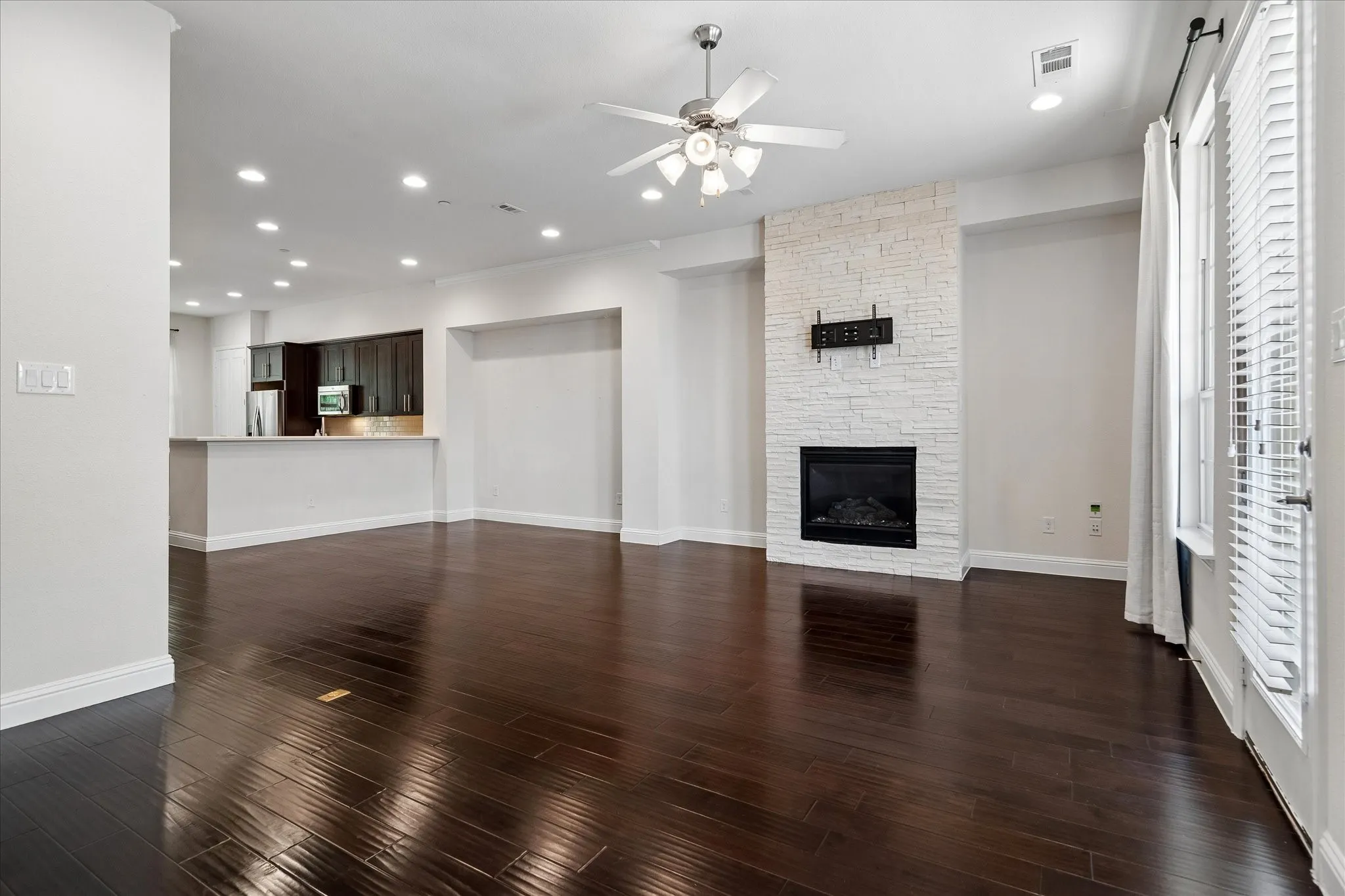 Dark wood-finish flooring, white painted walls, recessed lighting, ceiling fan, and a fireplace with a stacked stone-look surround