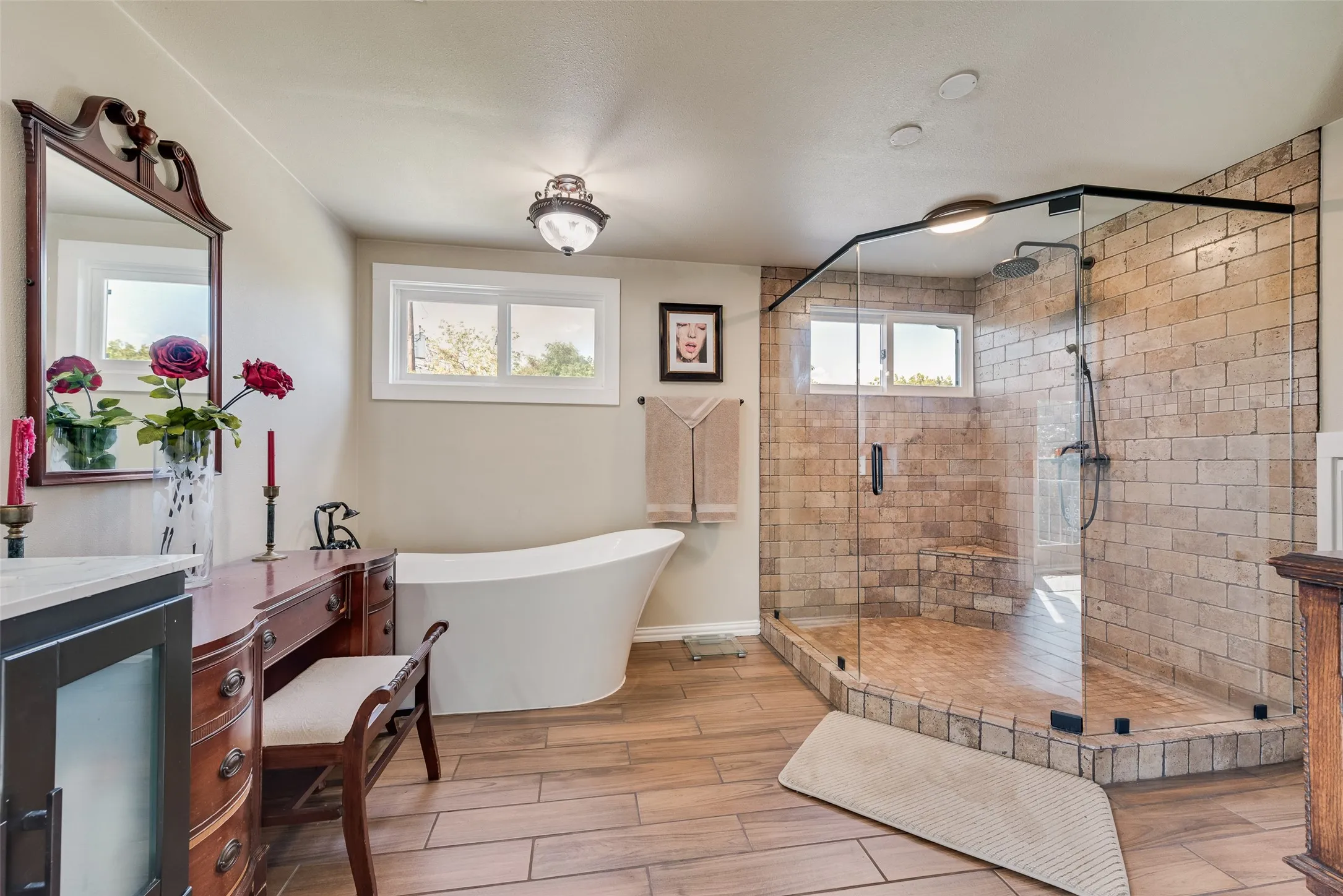 Freestanding soaking tub, walk-in shower with tile surround, glass shower enclosure, wood-finish flooring, and dual windows