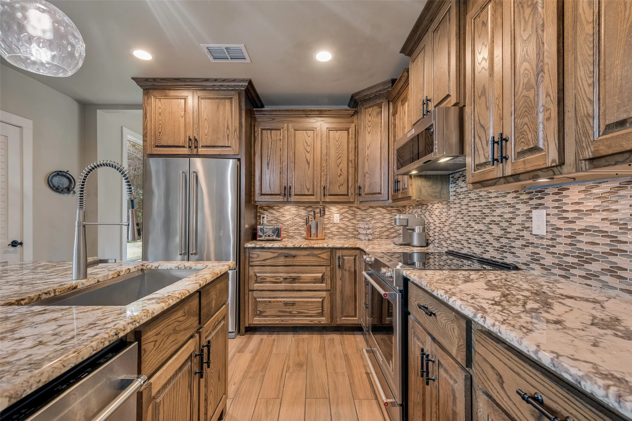 Wood-finish cabinetry with dark hardware, stone countertops, stainless steel appliances, mosaic tile backsplash, and wood-finish flooring