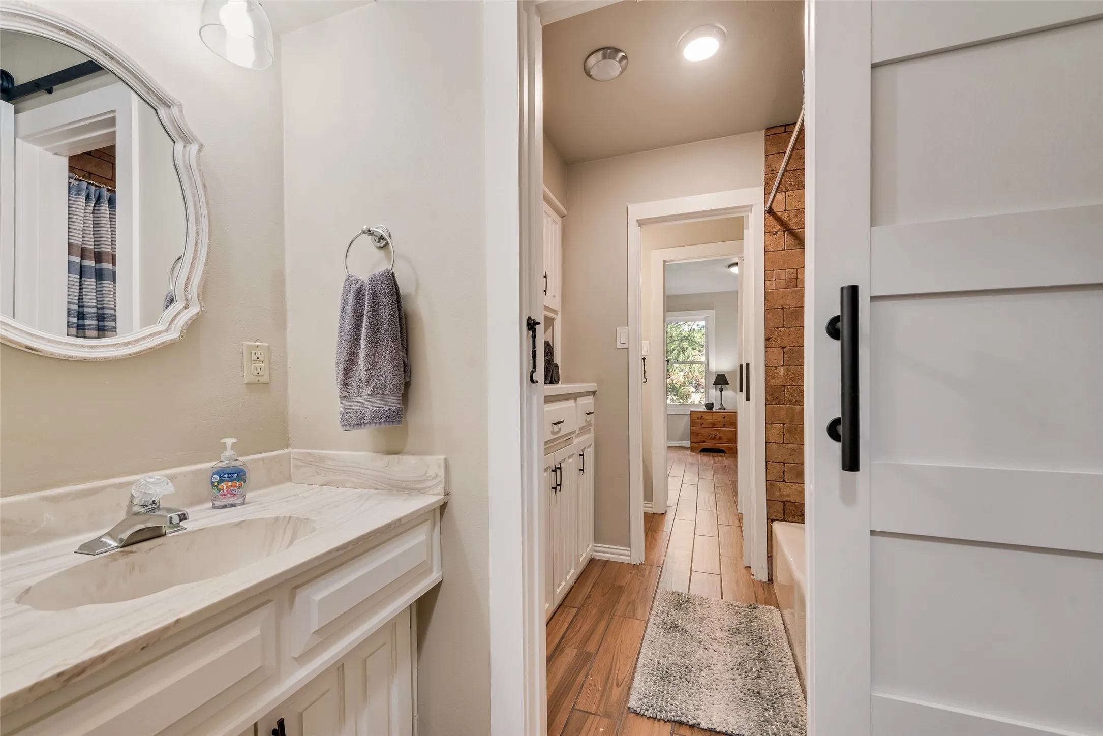 Vanity with integrated sink and chrome faucet, decorative framed mirror, white shaker-style cabinetry, white barn door with black hardware, and brick-look tile accent wall