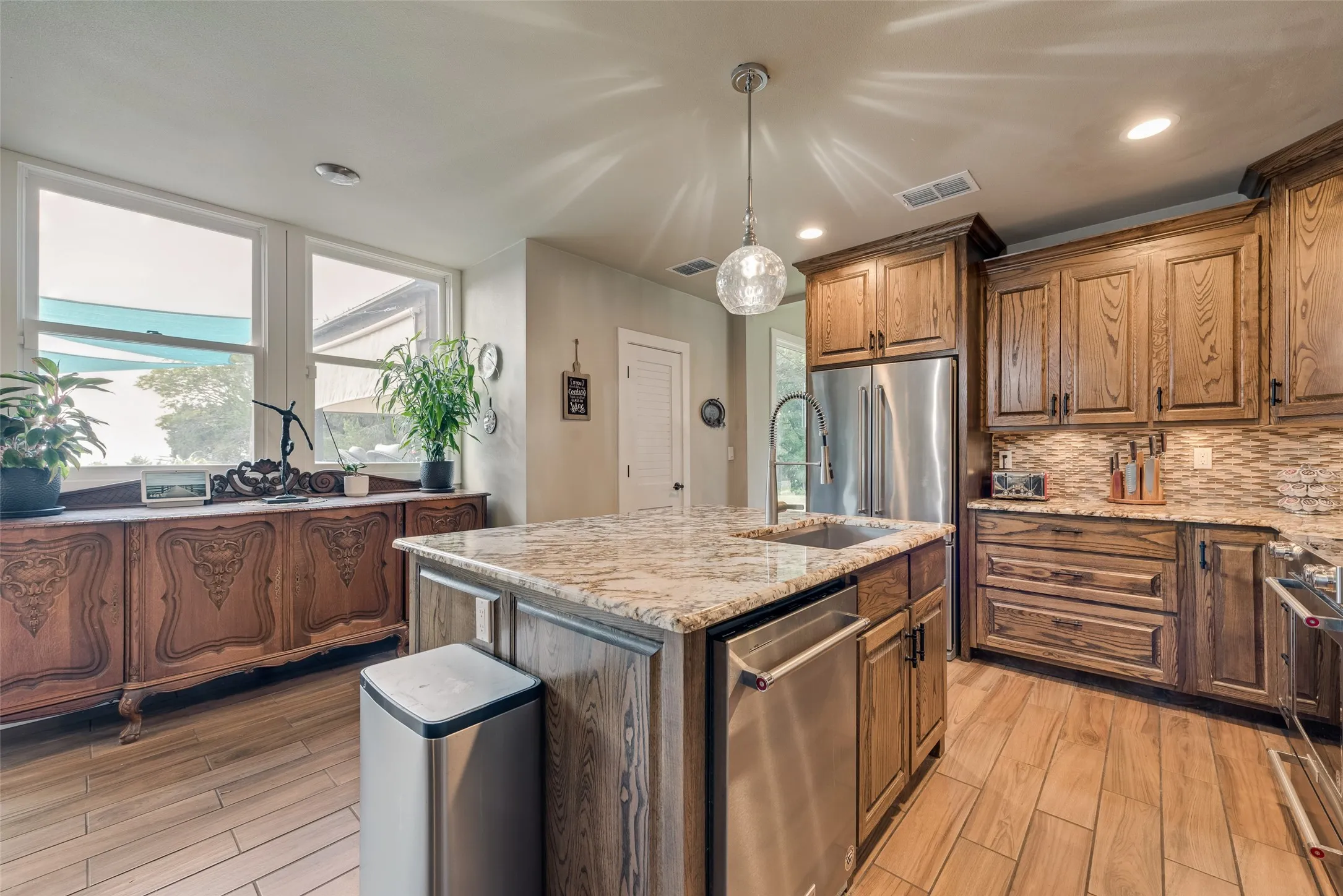 Kitchen featuring wood-finish flooring, a central island with a granite countertop, wood cabinetry, stainless steel appliances, and a mosaic tile backsplash