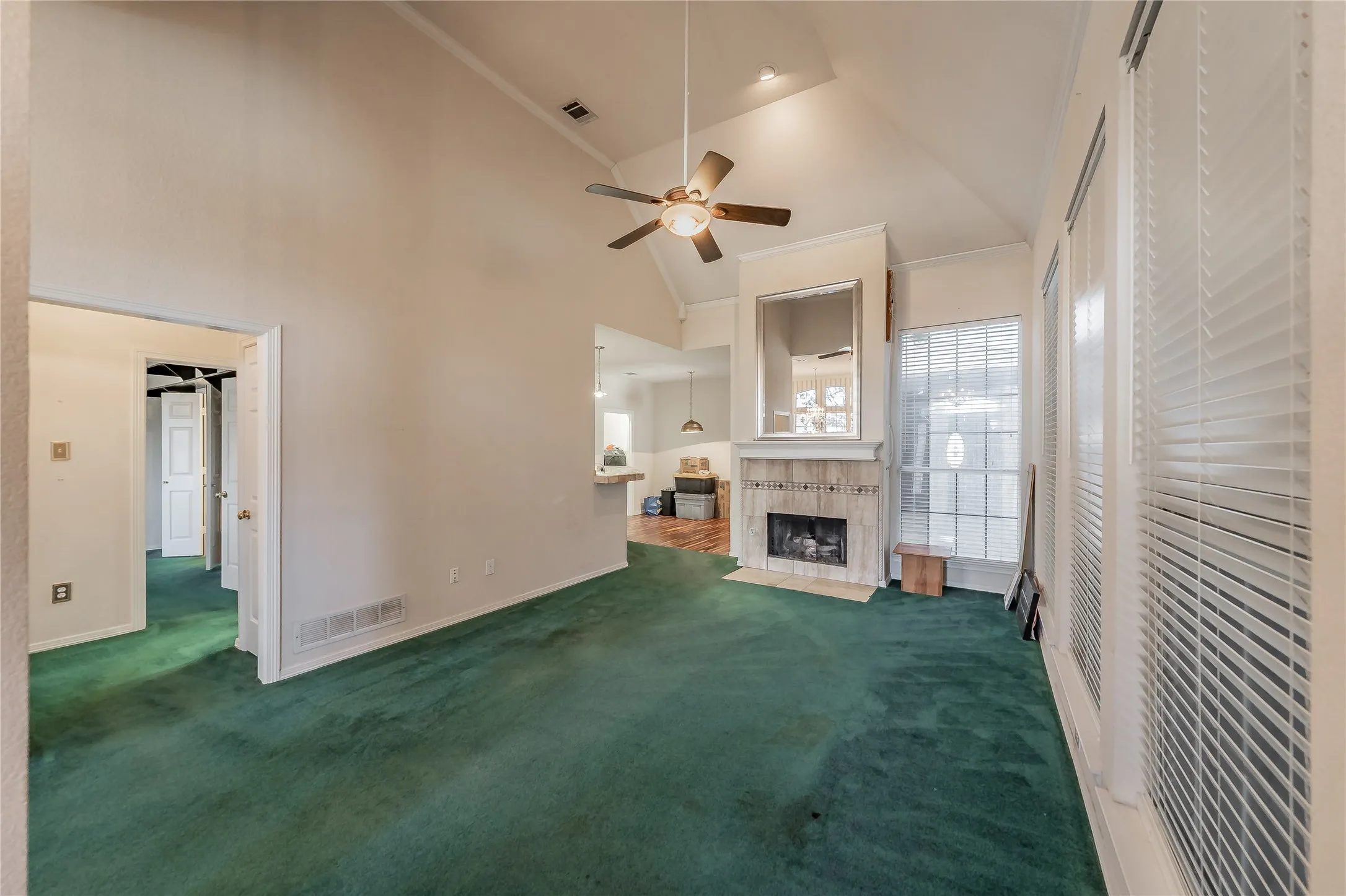 Vaulted ceiling with a ceiling fan, fireplace with a tile surround and mantel, and multiple windows with horizontal blinds