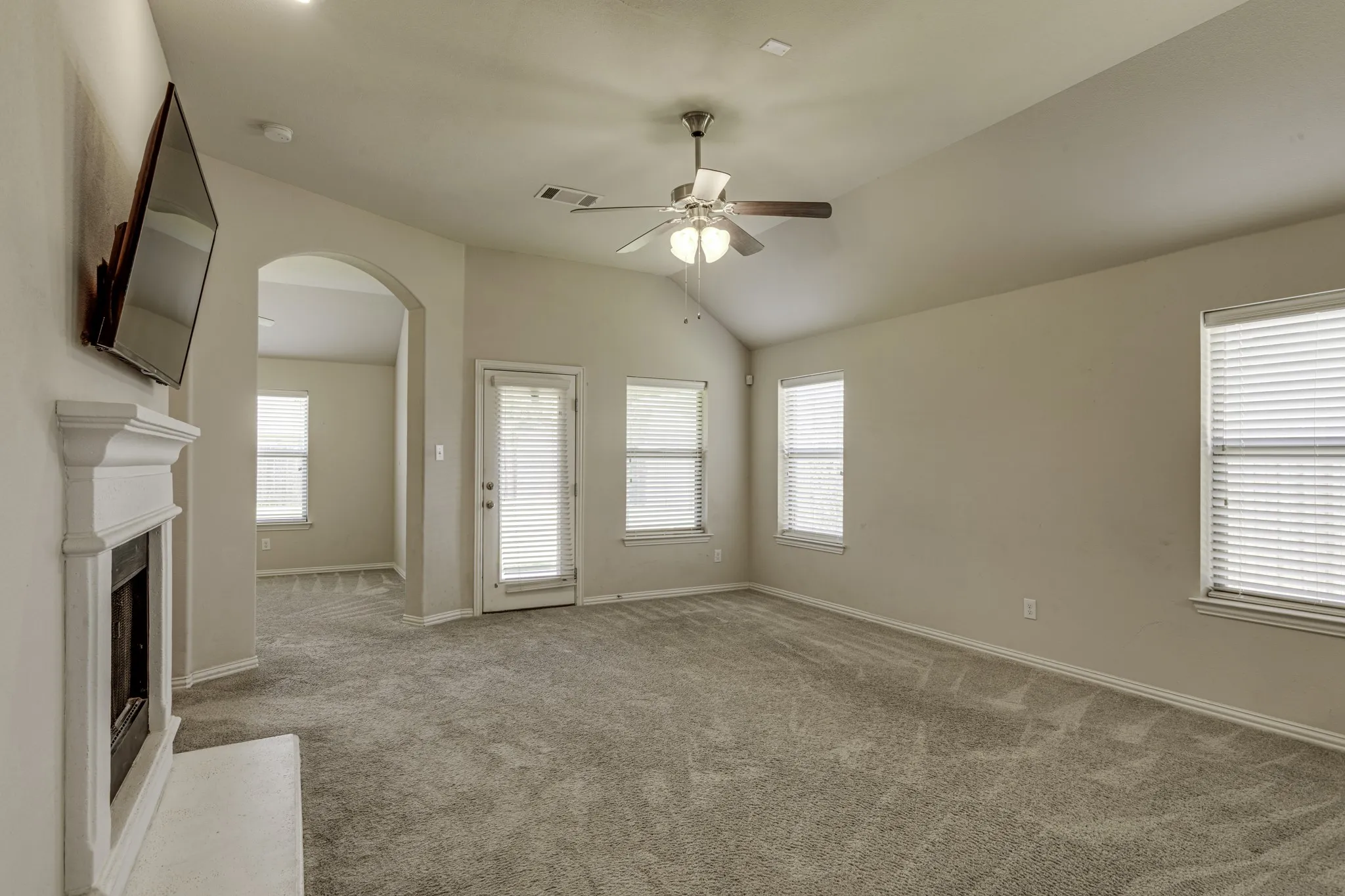 Carpeted living space featuring an arched doorway, a fireplace with a white mantel, multiple windows with blinds, a ceiling fan, and a light-colored vaulted ceiling.