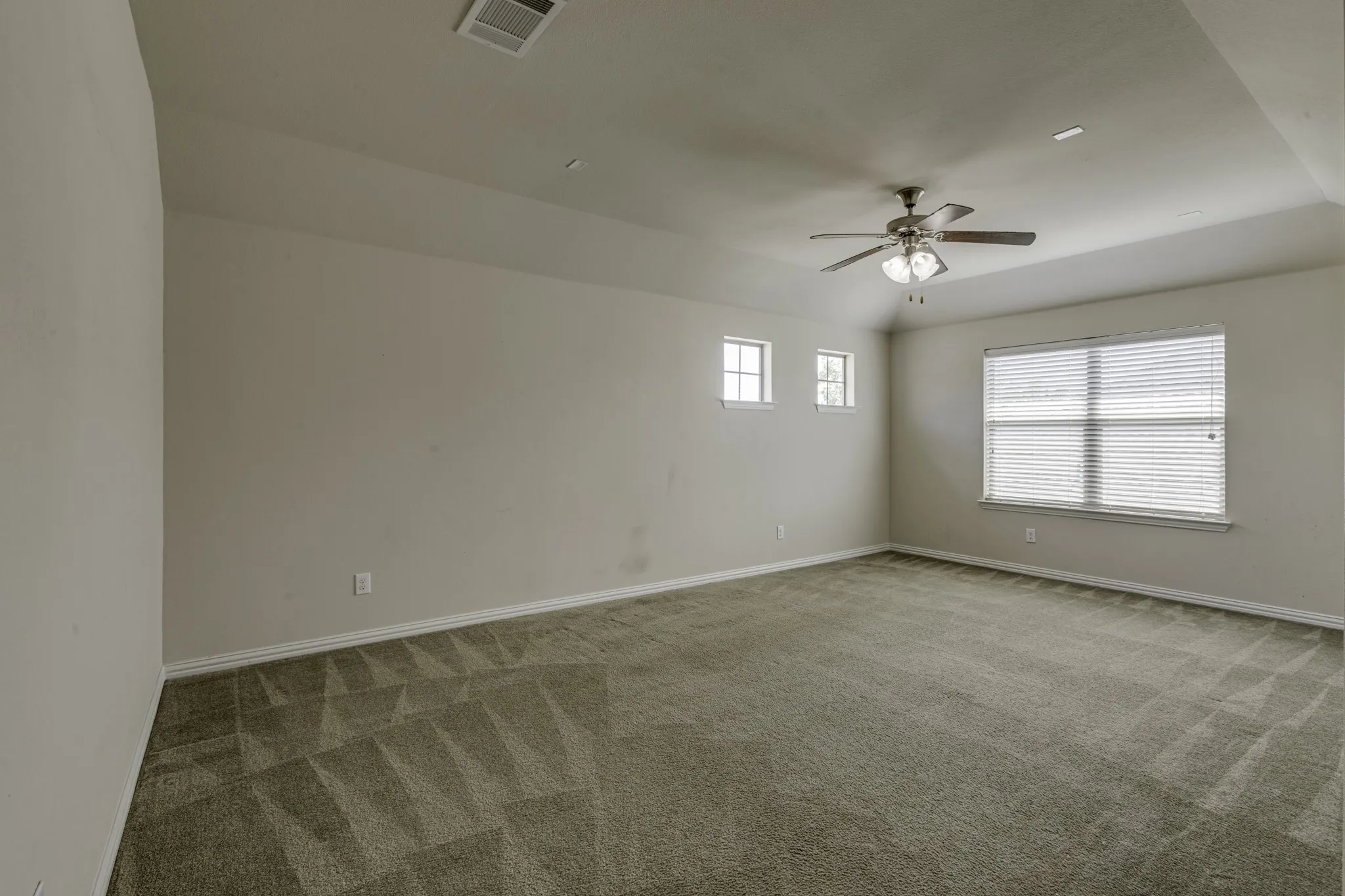 Spacious room featuring neutral carpeting, a tray ceiling, a ceiling fan with light fixture, and multiple windows with blinds