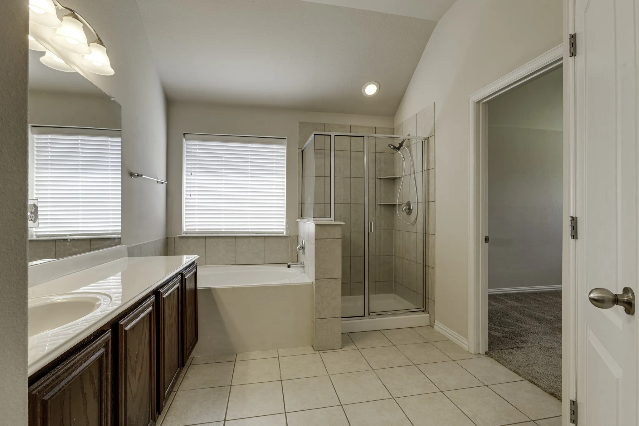 Dual sink vanity with dark wood-finish cabinetry, a drop-in soaking tub, a glass-enclosed shower with tiled walls, and light tile flooring.