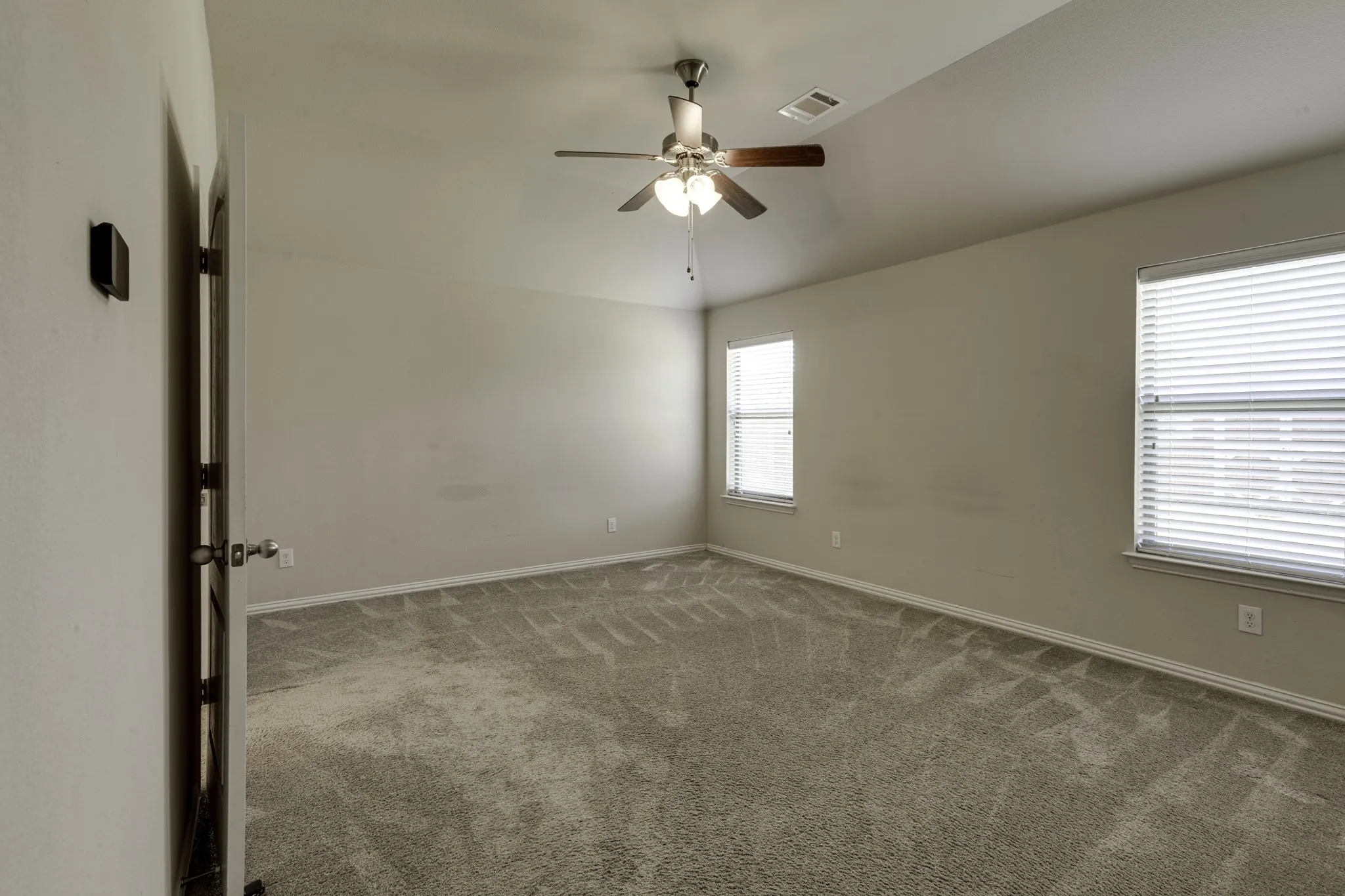 Master bedroom showcasing neutral wall finishes, plush carpeting throughout, ceiling fan with integrated lighting, and abundant natural light from dual windows with horizontal blinds.