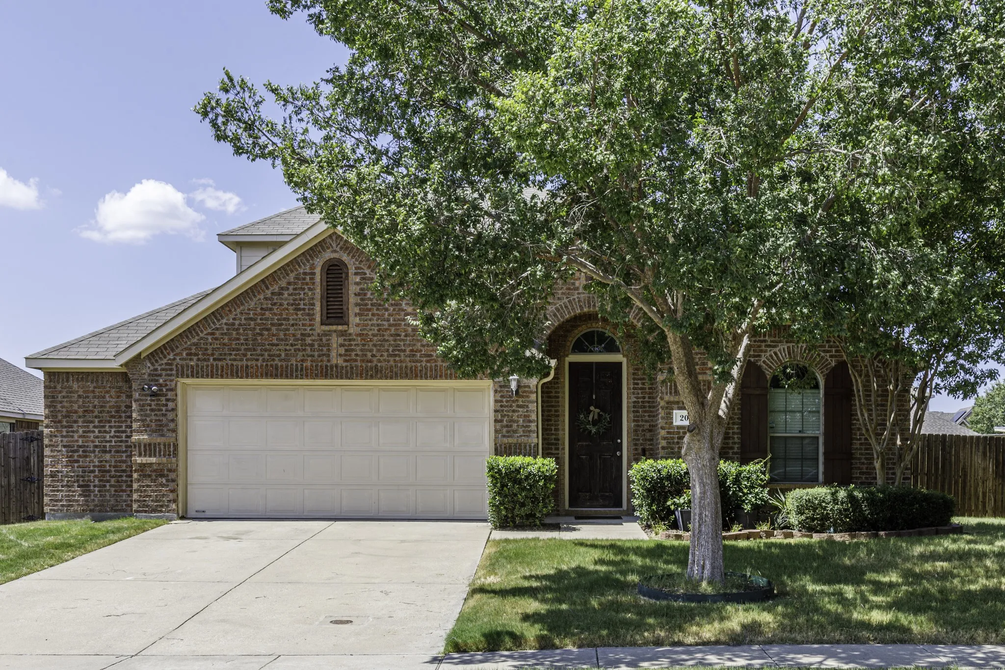 Brick Colonial home featuring a two-car garage with white panel doors, arched entry with dark wooden door, manicured front landscaping with mature trees, and a concrete driveway