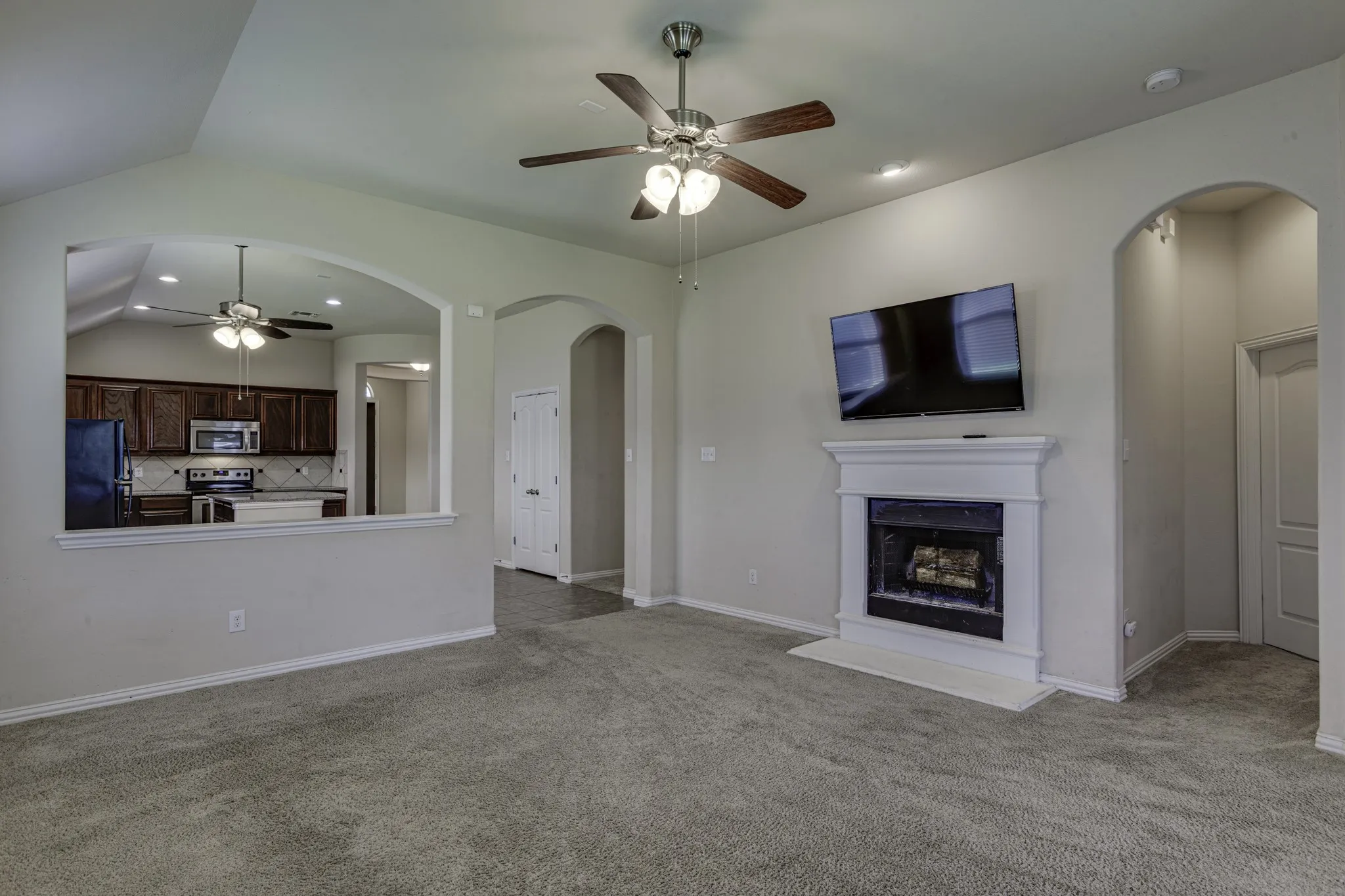 Spacious open-concept living area featuring neutral carpet flooring, ceiling fan with integrated lighting, and a gas fireplace with white surround and mantel