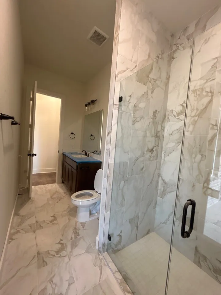 Glass-enclosed shower with tiled surround, wood-finish vanity with dual sinks, and patterned tile flooring