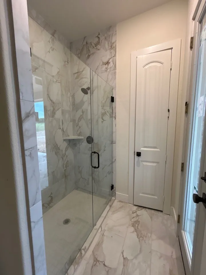 Walk-in shower with clear glass enclosure and matte black hardware, featuring marble-look wall tiling and a built-in shower shelf