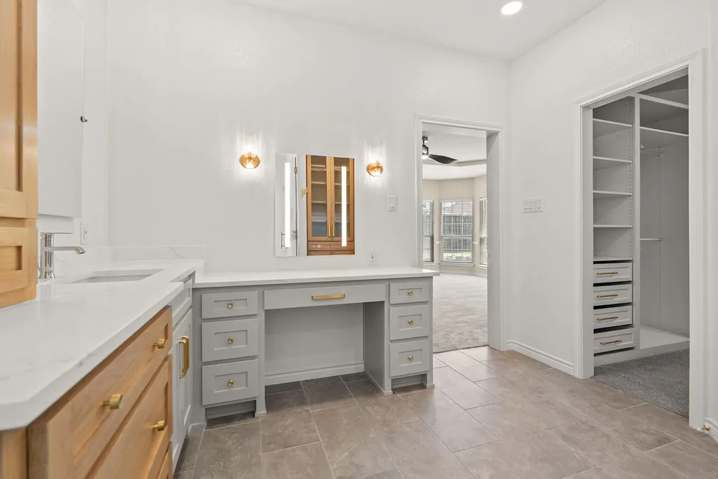 Light-toned wood-finish cabinetry, white countertop, undermount sink, gray vanity with gold-tone hardware, and built-in closet shelving