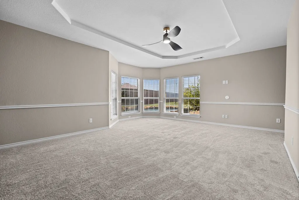 Tray ceiling with a ceiling fan, bay window with multiple windows, neutral tone carpet, and white trim molding
