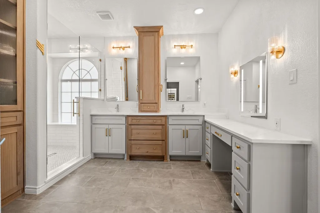 Dual vanities with quartz countertops, custom cabinetry, walk-in shower with glass enclosure, arched window, and integrated vanity lighting