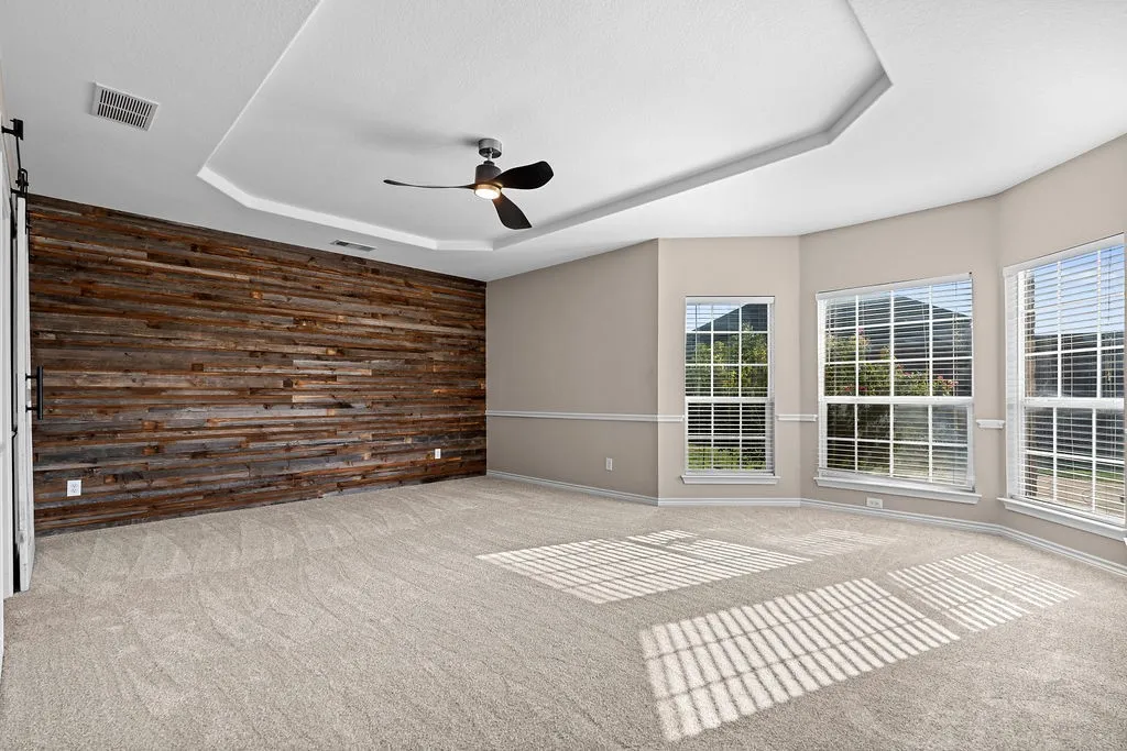 Tray ceiling, contemporary ceiling fan, accent wall with wood paneling, bay window, and light-toned carpeting