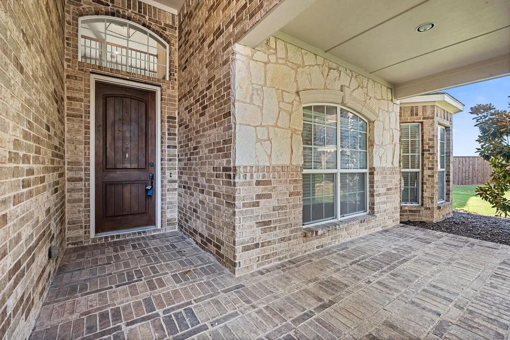 Brick and stone exterior with a prominent wooden entry door, arched transom window, and a covered porch featuring brick-style pavers