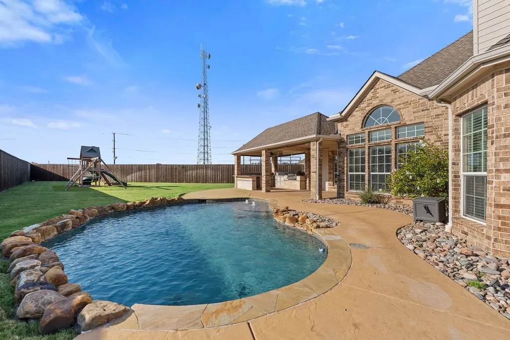Curved swimming pool with natural rock accents, stamped concrete patio, covered outdoor kitchen, and brick exterior