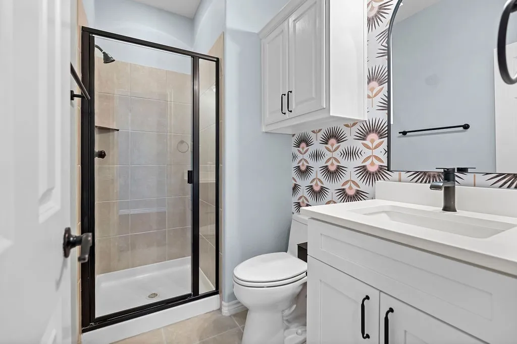 Walk-in shower with a black-framed glass enclosure, white vanity with an integrated sink, black-finish faucet, and patterned wallpaper accent