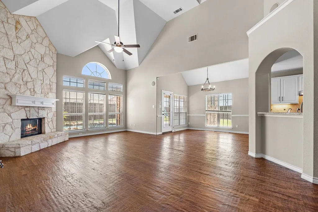 Vaulted ceilings, a stone-clad fireplace with mantel, expansive window array, wood-finish flooring, and an arched entry to the kitchen with white cabinetry