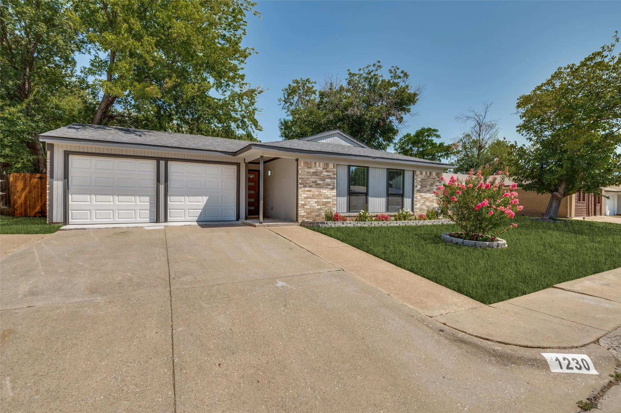Modern brick ranch featuring a two-car garage with contemporary paneled doors, manicured front lawn with mature landscaping, blooming crepe myrtle accent planting, concrete driveway, and a gabled roofline with updated architectural detailing.