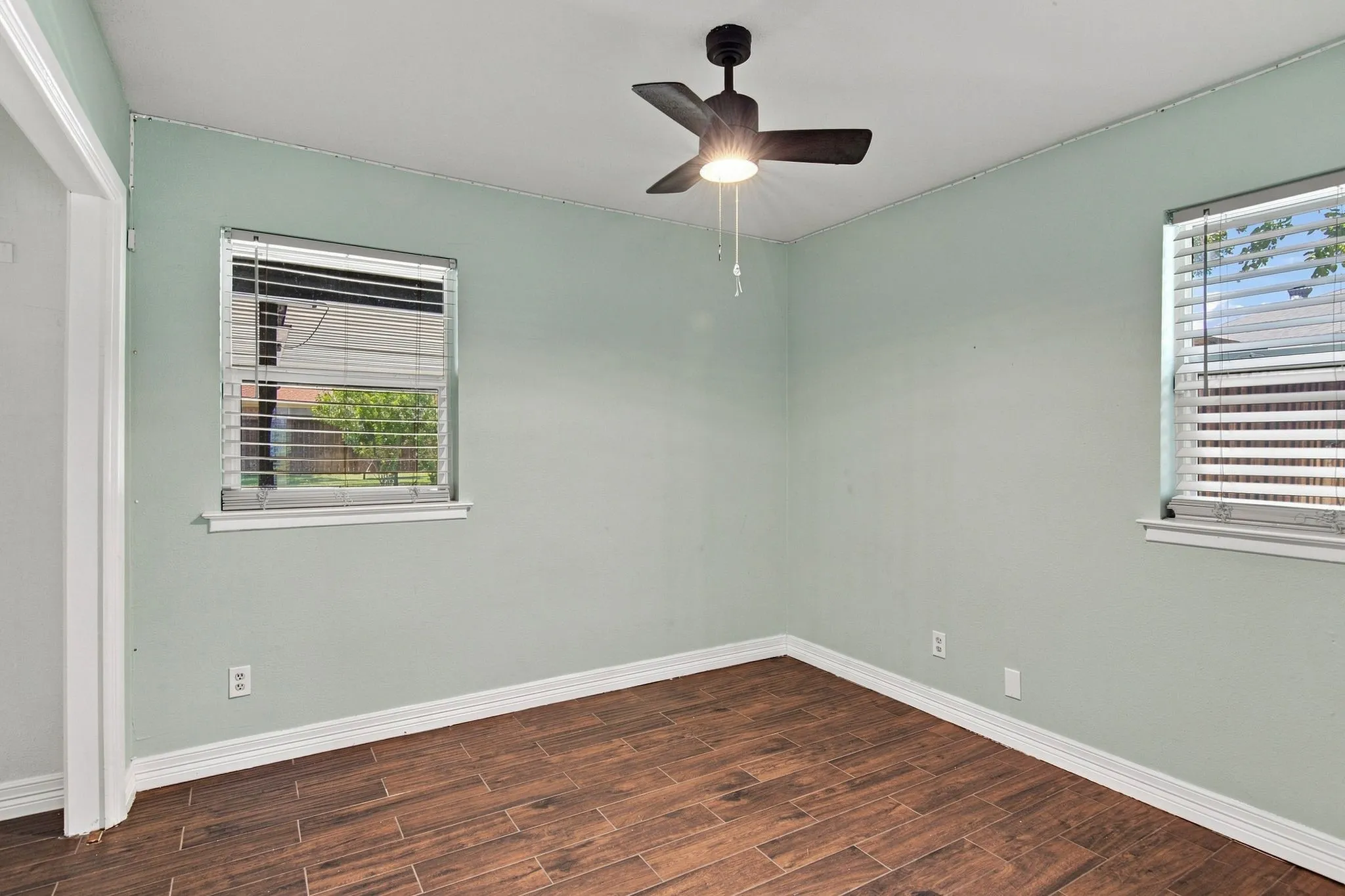 Wood-finish flooring, two windows with blinds, a ceiling fan with integrated lighting, and white trim