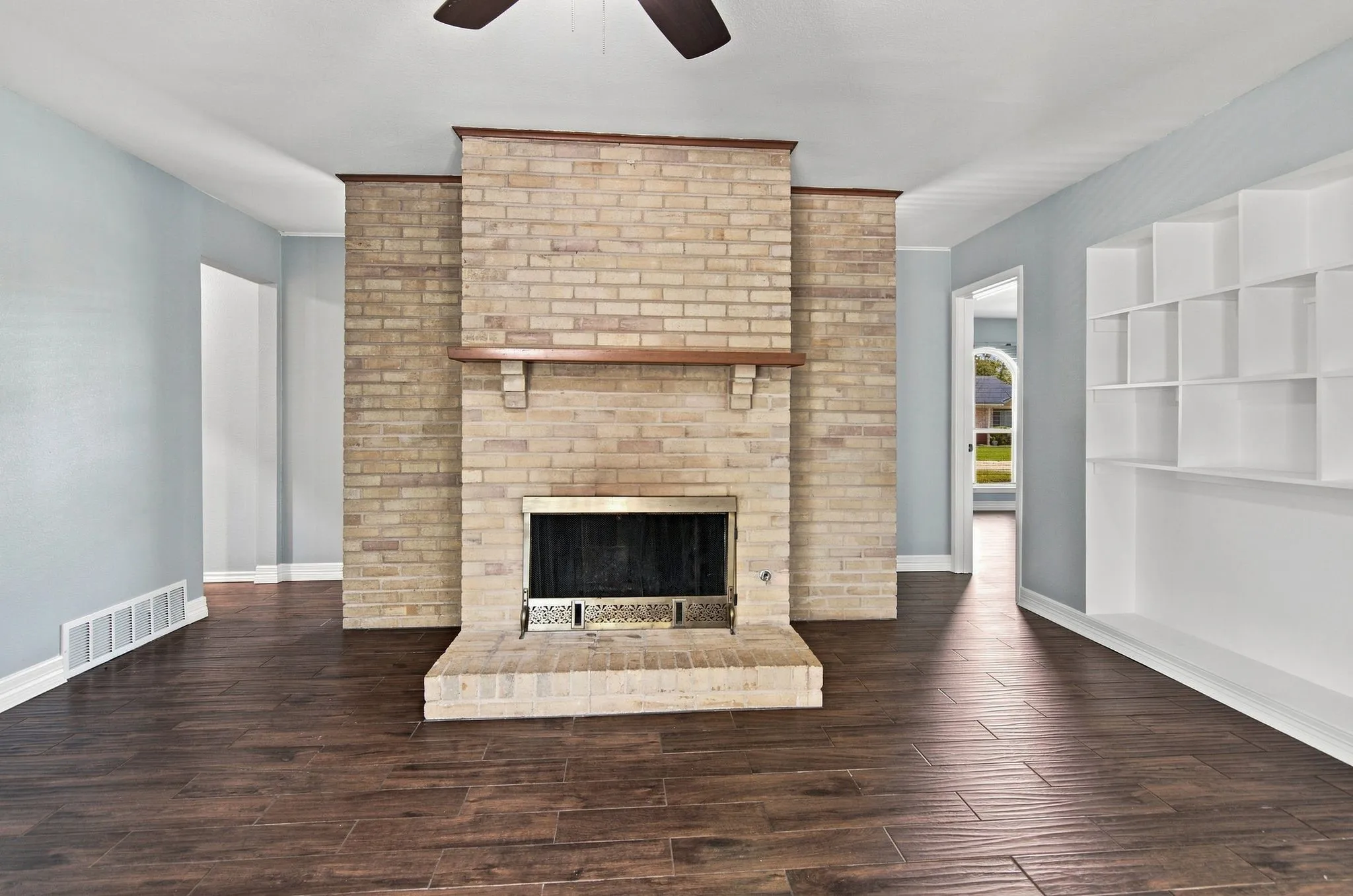 Brick gas fireplace with wood-finish tile flooring, built-in shelving, and a ceiling fan