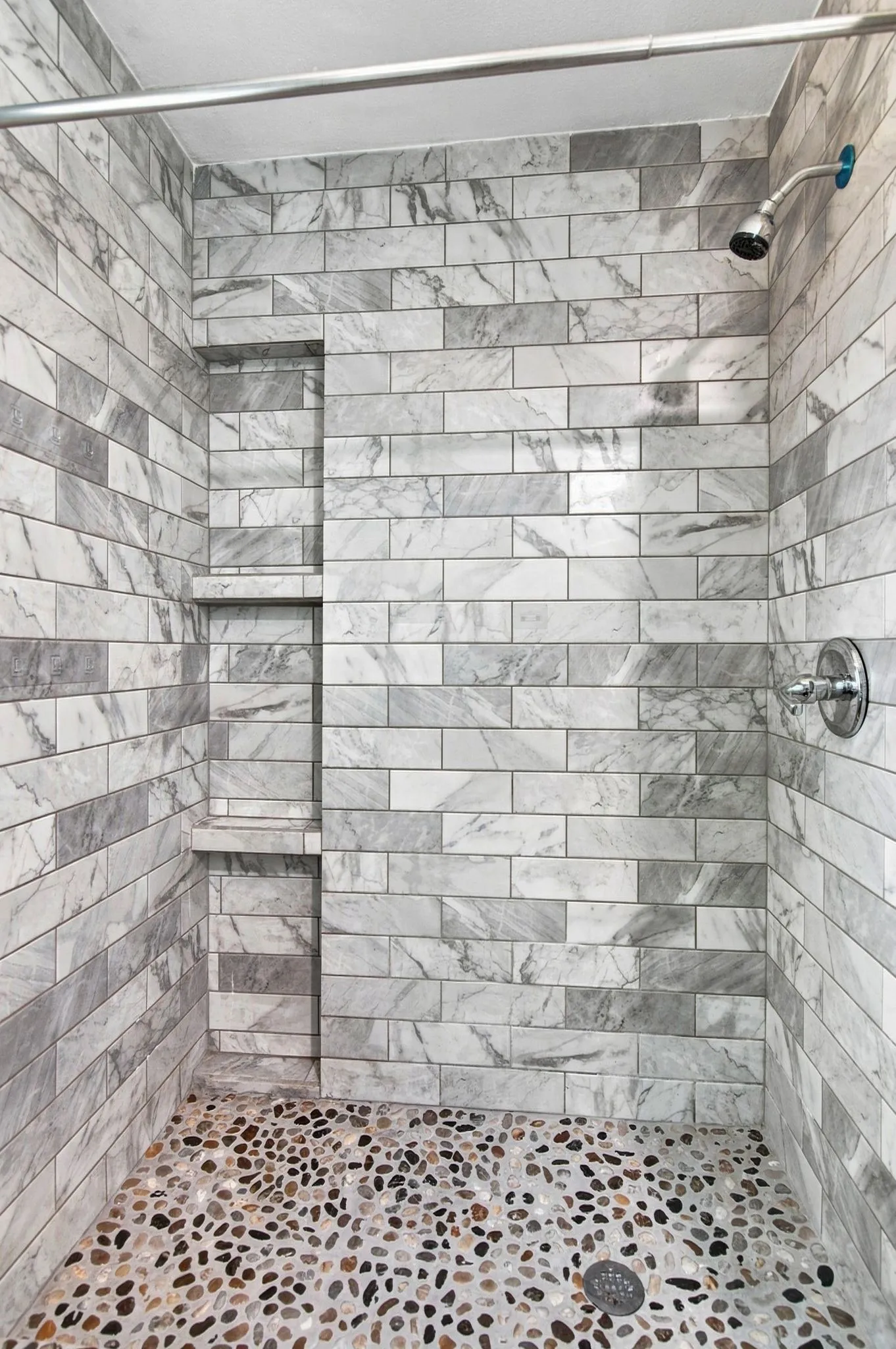 Shower stall featuring subway-style wall tiling, a built-in shower niche with two shelves, a pebble-finish floor, and a chrome shower head and controls