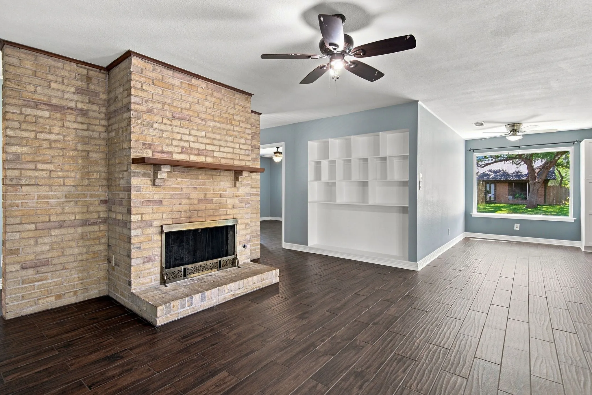 Brick fireplace with a wood mantle, wood-finish flooring, built-in shelving, and a ceiling fan