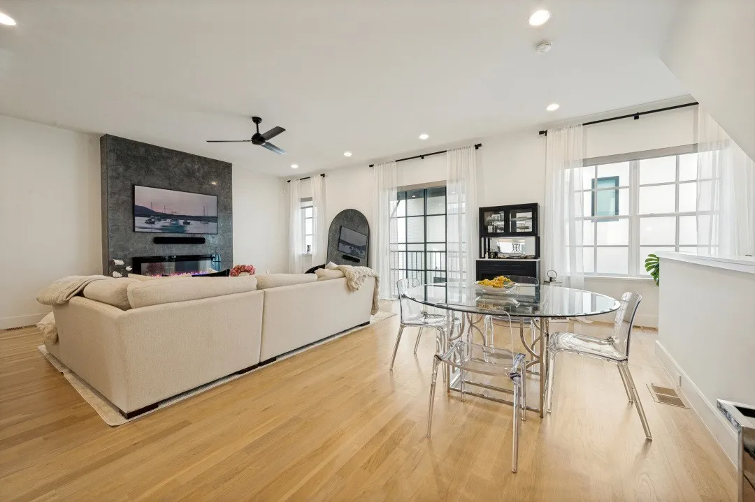 Open-concept living space featuring warm wood-finish flooring throughout, a cream upholstered sofa with modern lines, recessed ceiling lighting, and a ceiling fan