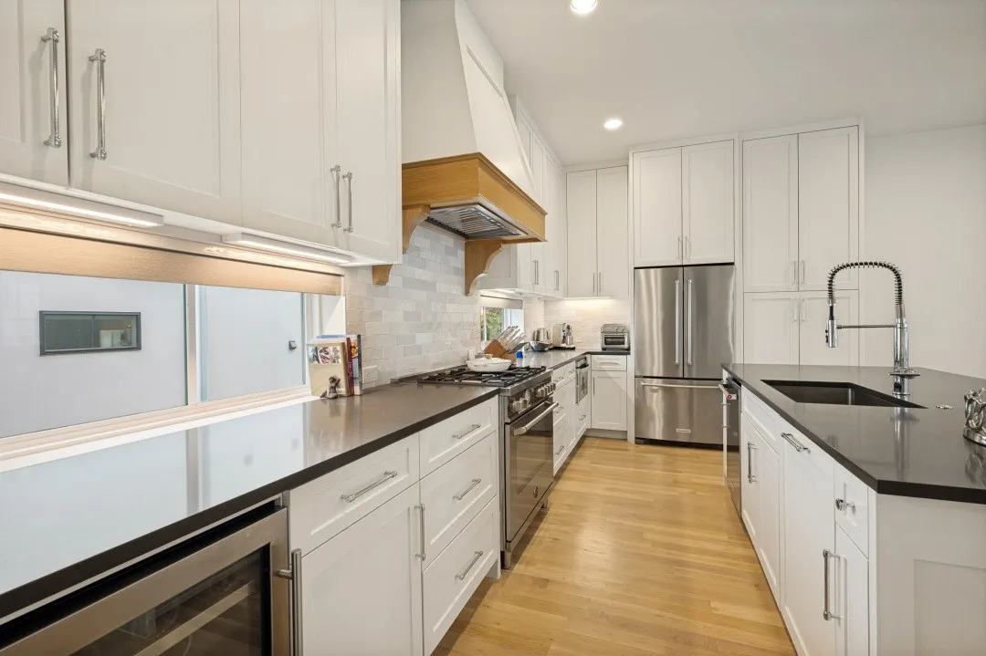 White cabinetry with brushed nickel hardware, stainless steel appliances, dark countertops, wood-finish flooring, and a kitchen island with an integrated sink