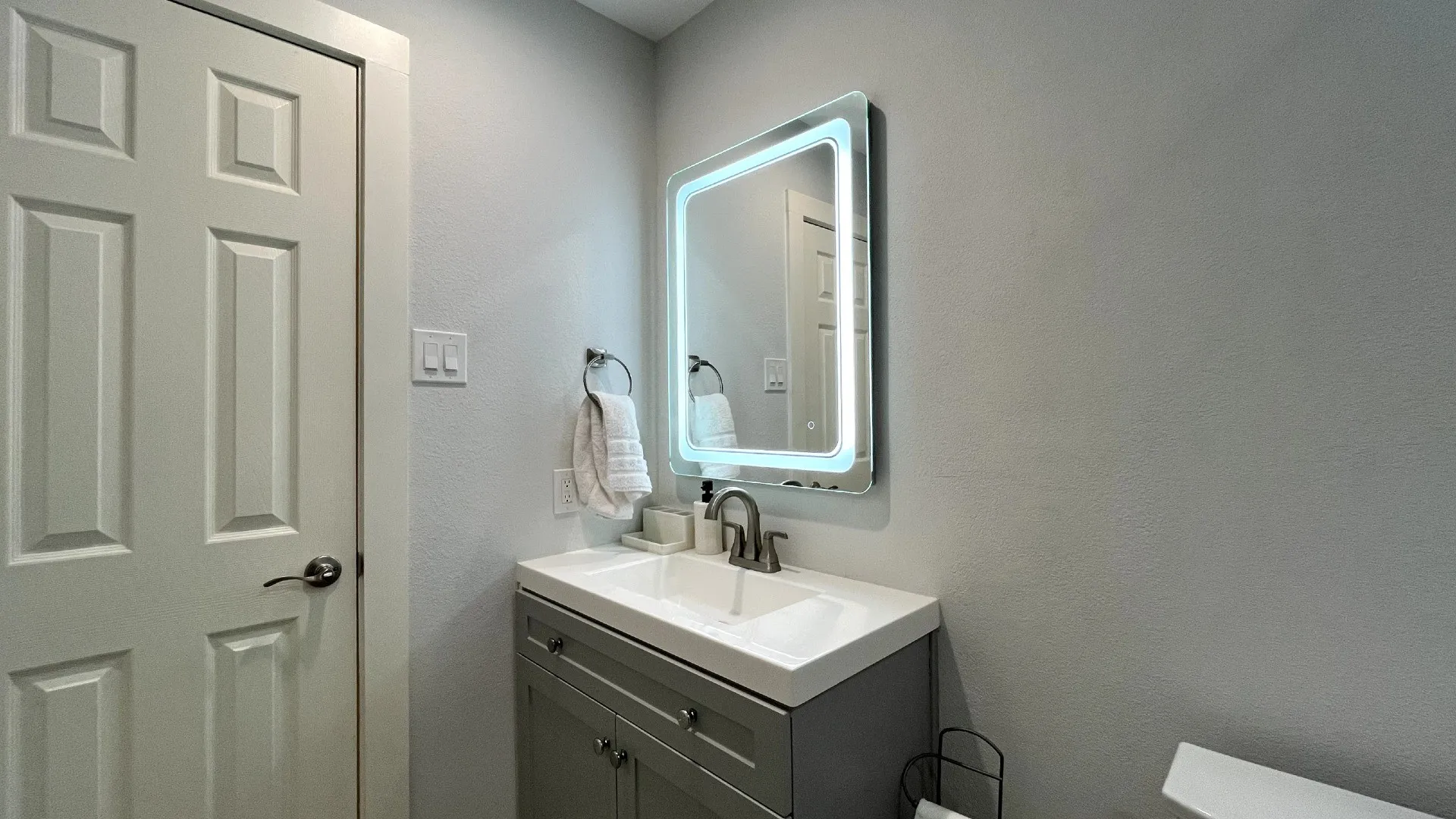 Backlit vanity mirror, single basin sink, gray vanity cabinetry, and a chrome faucet