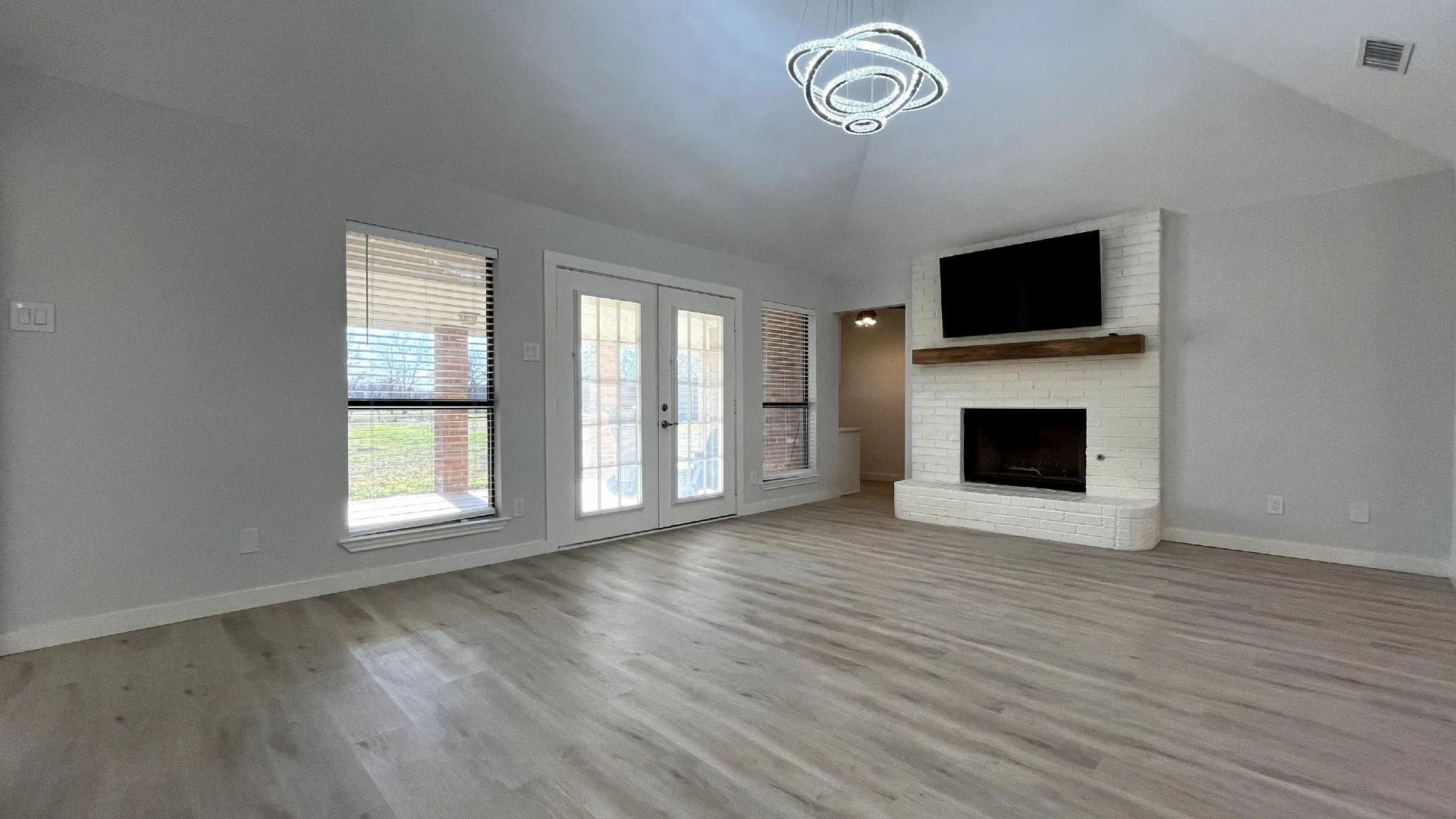 Spacious living area featuring light wood-finish flooring, a white brick fireplace with a wood mantle, and double glass-paneled doors