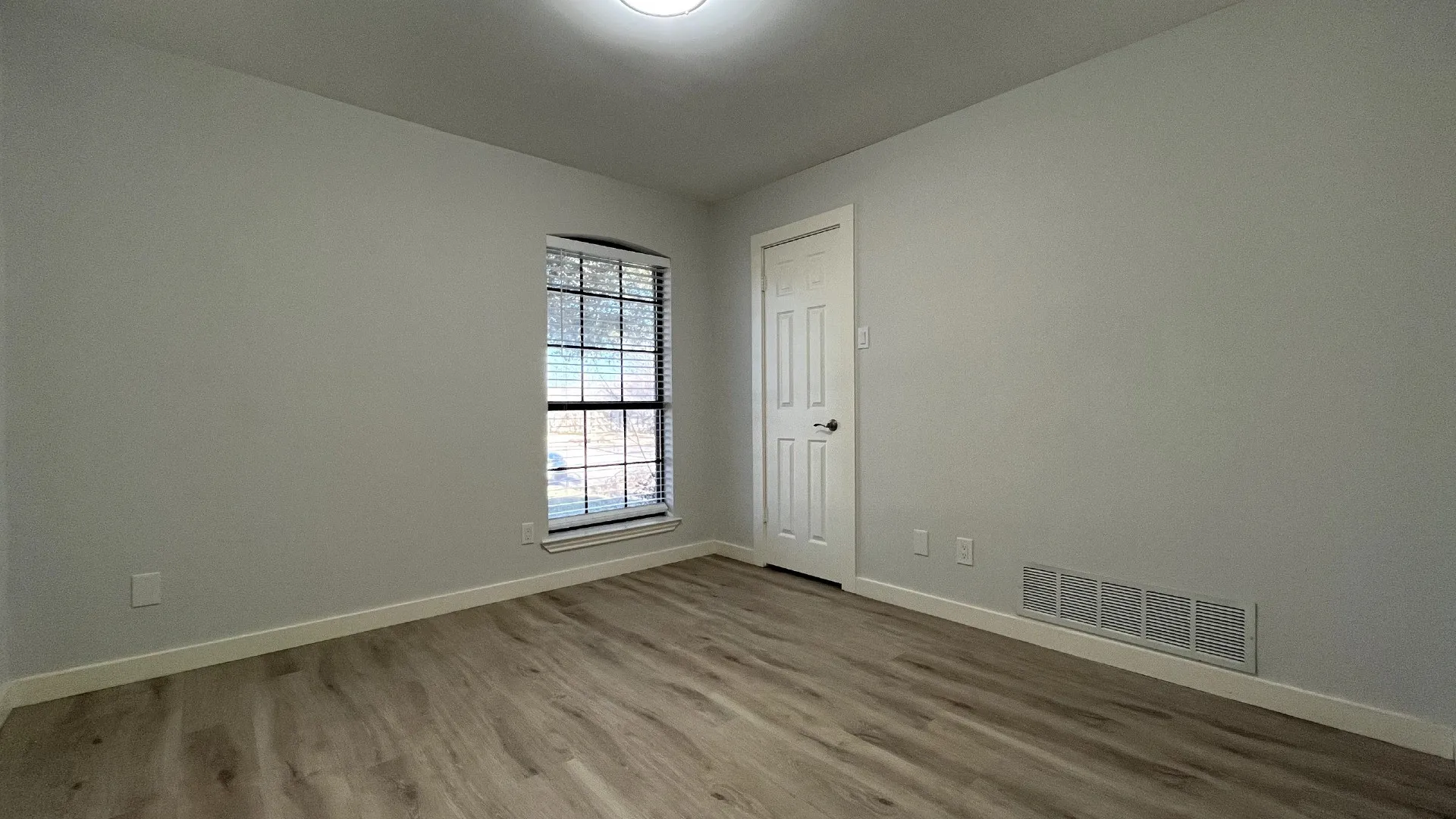Wood-finish flooring, neutral wall tones, window with blinds, paneled door, and overhead lighting